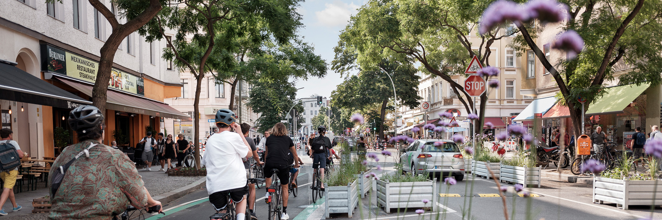 Eine belebte Straße auf der ein Radweg mit mehreren Radfahrenden von der Straße durch mit Blumen bepflanzte Hochbeete getrennt ist.