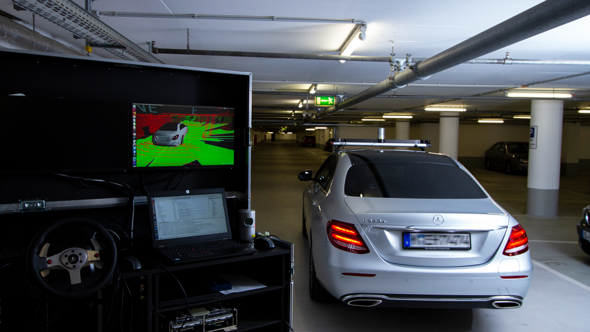 Eine mobile Kontrollstation für Teleoperiertes Fahren mit 2 Bildschirmen und einem Lenkrad in einer Tiefgarage mit einem silbernen Fahrzeug im Hintergrund.
