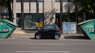 Ein kleines Auto parkt am Straßenrand zwischen zwei großen Containern für Bauschutt. Im Hintergrund sind auf den Gehweg ein Biefeinwurfkasten und ein Stromverteilerkasten zu sehen.
