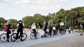 10 Personen auf Fahrrädern und eine Person auf einem Elektroroller warten an einer Ampel an einer großen Kreuzung auf dem Radweg