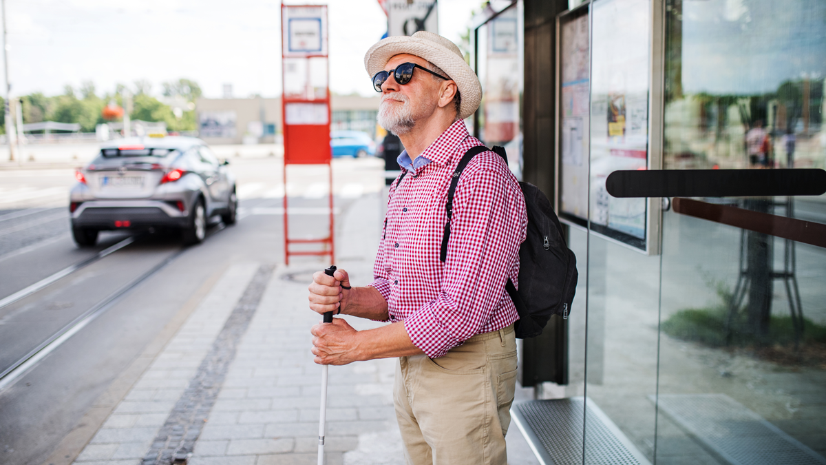 ASCT, News: Stadt Soest, barrierefreie automatisierte Busse Ein blinder Mann mit Hut, Sonnenbrille und Langstock steht an einer Bushaltestelle.