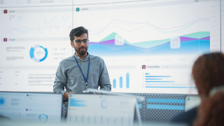 Male Indian Computer Scientist With Tablet Giving Presentation to Diverse Team Of Data Analysts In Front Of Big Digital Screen In Monitoring Room. Colleagues Listening To Training On Automation.