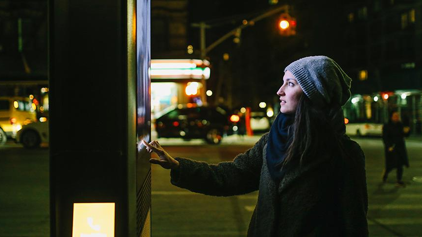 DPS, News: Anwenderzentrum für KI in Kommunen Eine Frau in Wintermantel und Mütze bedient eine digitale Litfaßsäule in einer Stadt bei Nacht