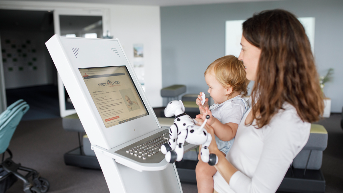 Eine Mutter steht mit ihrem Kind vor einem Terminal zur Beantragung von Kindergeld.