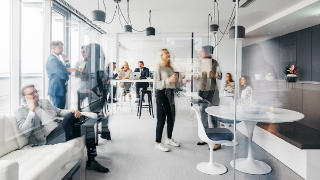 A photo showing a daily routine at the office. Some are standing up having conversations, some are sitting down chatting. They are smartly dressed. The office is spacious, modern and has large windows. Horizontal daylight indoor photo.