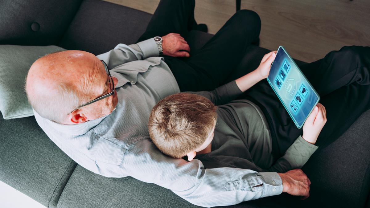 DPS, Projekt, Vorschaubild: Quartier Zukunft Grandfather and grandchild sit in a sofa in a living room. The boy holds on to a digital tablet, and they both look at the screen. The screen shows a fictive smart home app, which can control items in the house like the washing machine and door locks.