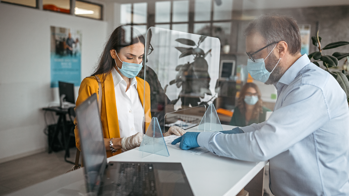DPS, News: Digitalisierung der Verwaltung Corona-Pandemie Eine Frau wird durch einen Verwaltungsmitarbeiter an einem Empfangstresen beraten. Beide tragen einen Mund-Nasen-Schutz, Handschuhe und sind durch eine Plexiglasscheibe getrennt.