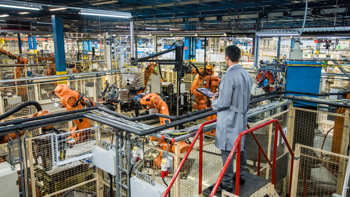DPS, News, Projekt: XMANAI Mann mit Tablett und grauem Arbeitskittel steht in Fabrikhalle mit Robotern