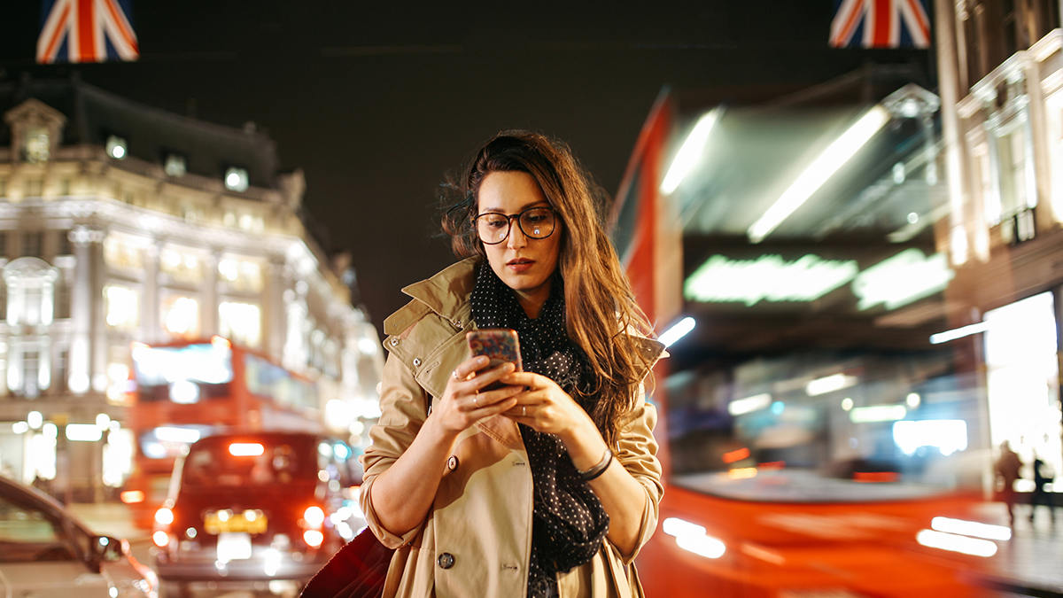 DPS, Projekte: iCity Vintage toned portrait of a young woman, wearing a trench coat, walking on the busy streets of London in the evening. Going home after work, texting on her cellphone, trying to find Uber or a cab.