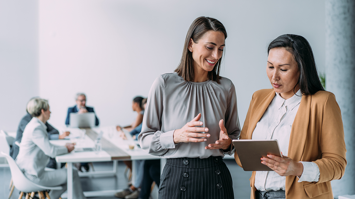 Shot of two female coworkers having a discussion in modern office. Businesswomen in meeting using digital tablet and discussing business strategy. Confident business people working together in the office. Corporate business persons discussing new project and sharing ideas in the workplace. Successful businesswomen standing with their colleagues working in background.