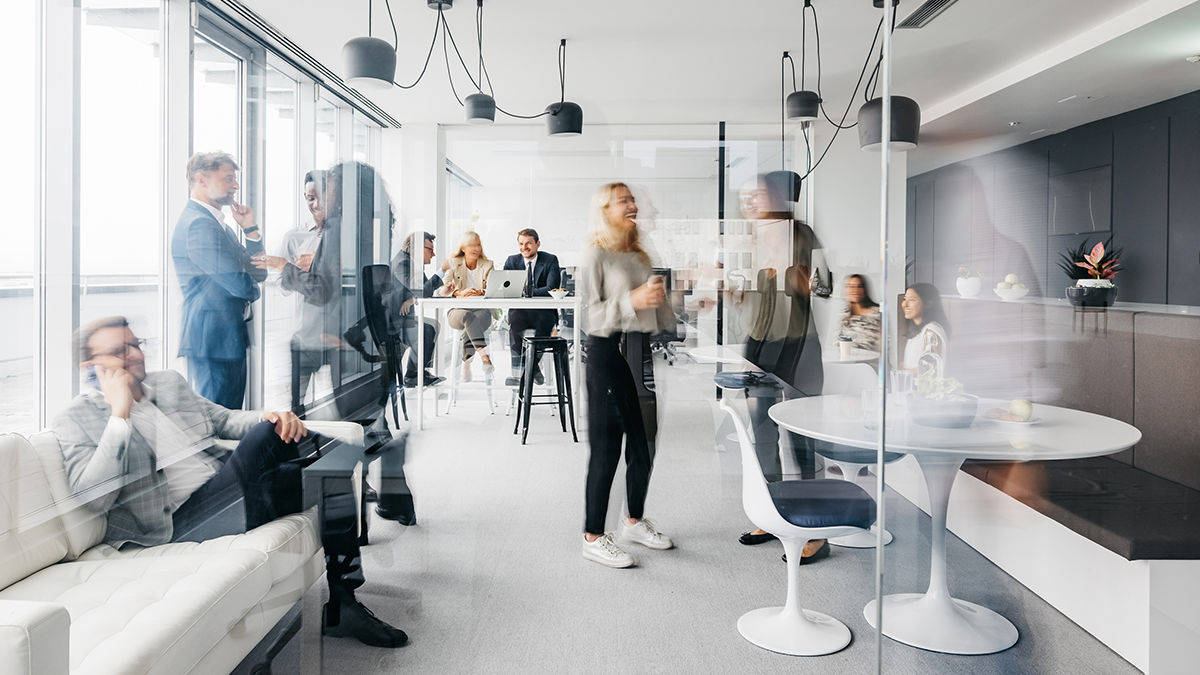 A photo showing a daily routine at the office. Some are standing up having conversations, some are sitting down chatting. They are smartly dressed. The office is spacious, modern and has large windows. Horizontal daylight indoor photo.