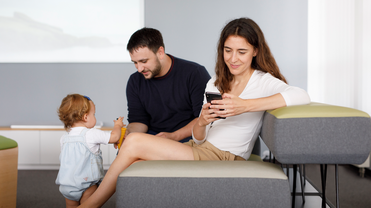 KI-Sprachassistent für Bürgerdienste: Frau sitzt auf einem Sofa und nutzt Bürgerdienste auf dem Smartphone, im Hintergrund spielt der Mann mit dem Kleinkind