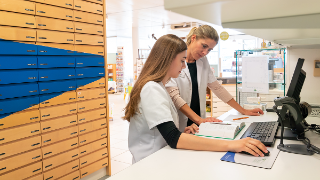 A female blonde pharmacist helps and assists her young aprrentice trainee during studies of medical books