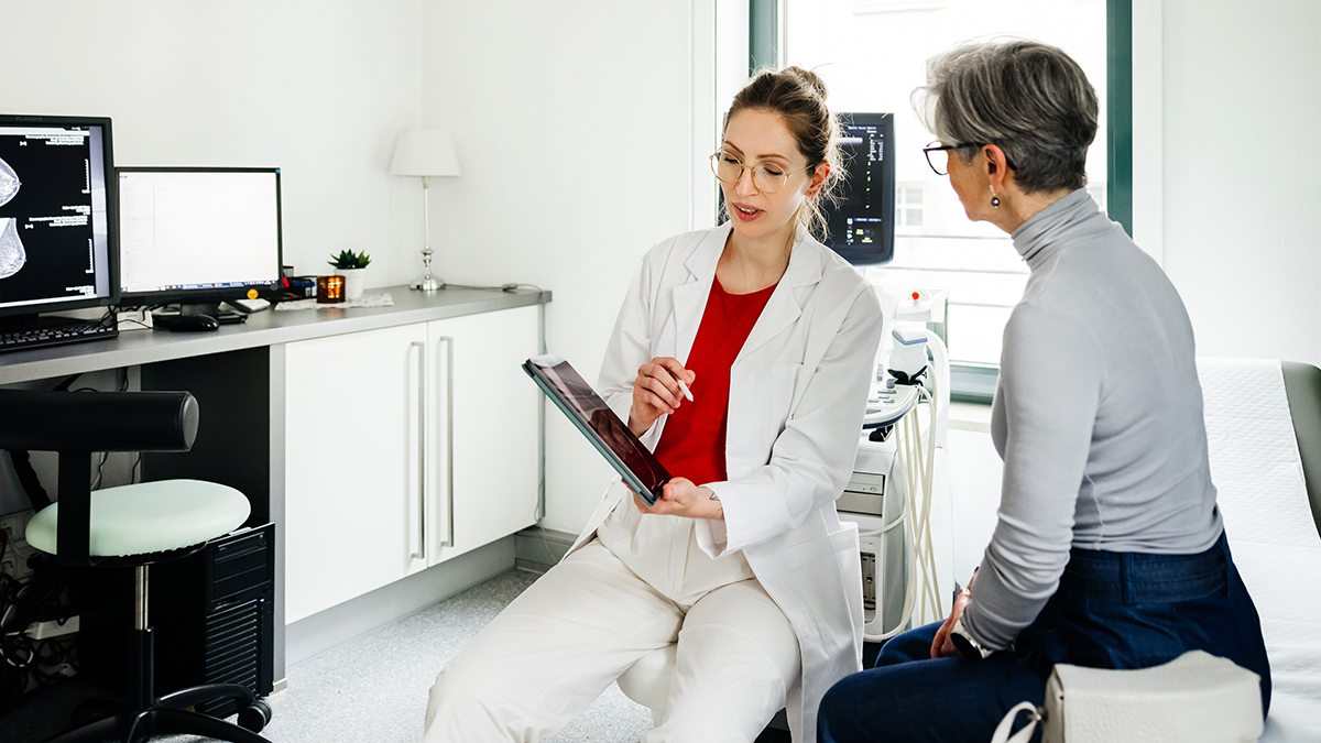 Young Female doctor is holding a digital tablet and explaining medical test results to  senior patient seated on the examination bed. The room is equipped with medical devices and a computer workstation.