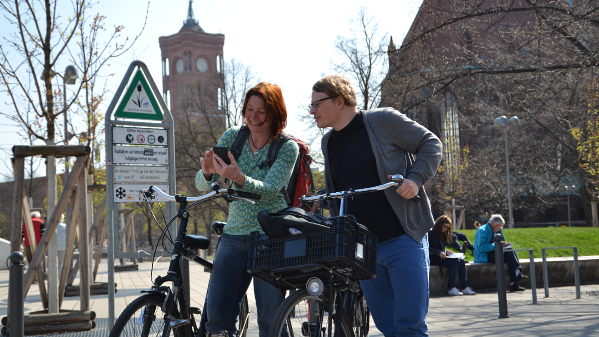 Zwei Radfahrende stehen auf dem Alexanderplatz mit dem Roten Rathaus im Hintergrund und schauen auf ein Smartphone