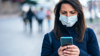 Porträt einer jungen Frau auf der Straße, die eine Gesichtsschutzmaske zur Vorbeugung von Coronavirus und Anti-Smog trägt und ein Smartphone benutzt