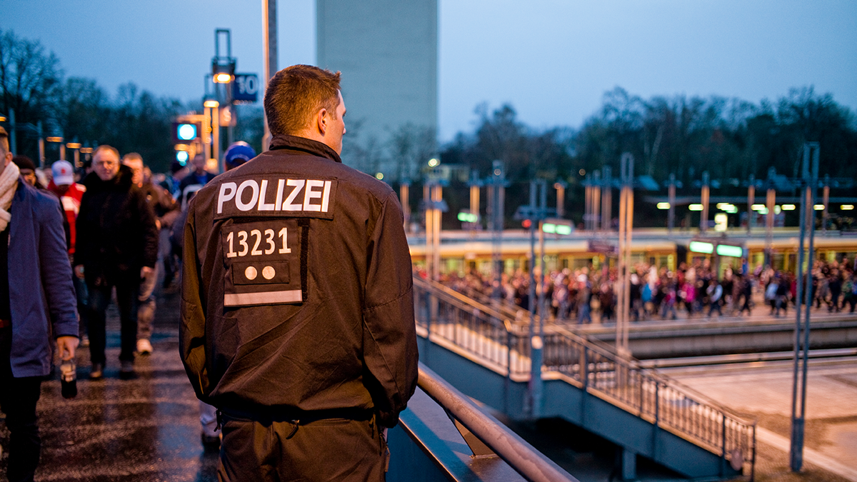 Ein Polizist steht auf einer Brücke mit dem Rücken zur Kamera, während ihm eine große Menschengruppe aus Richtung eines S-Bahngleises entgegenkommt.