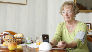 Seniorin misst am Frühstückstisch ihren Blutdruck mit einer Manschette, die die Werte an ein angeschlossenes Smartphone überträgt.