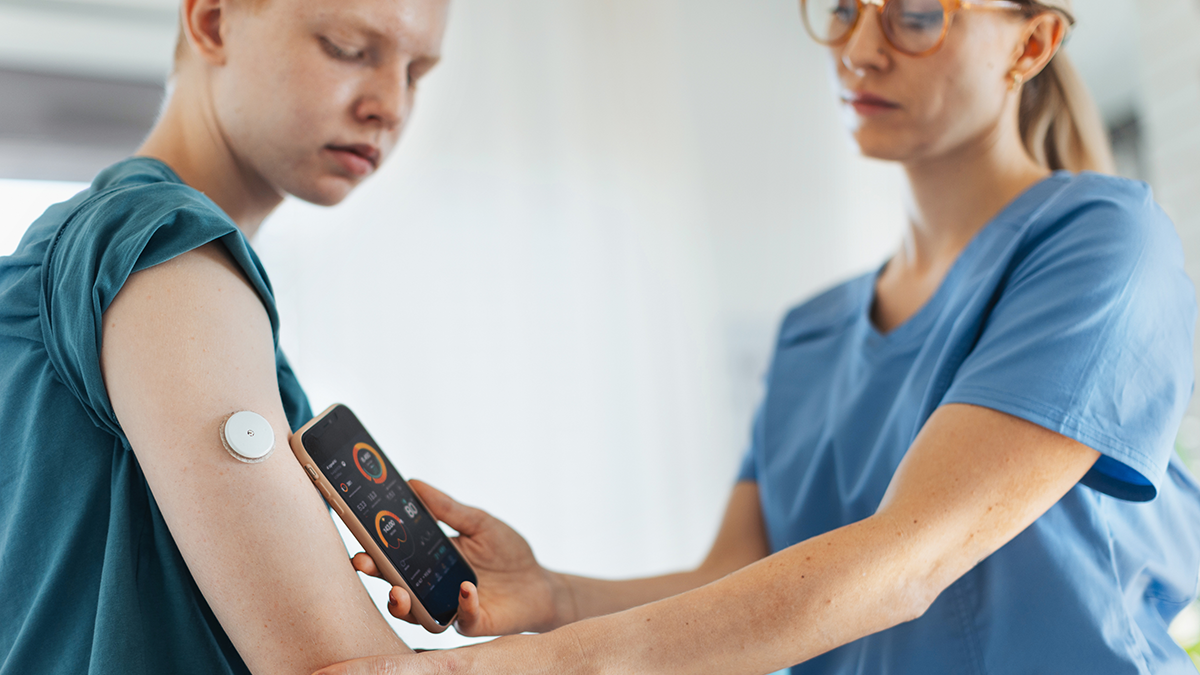 Diabetologist doctor connecting continuous glucose monitor sensor to smartphone, checking teenage boy's glucose data in the real time. Endocrinologist taking care of teenage diabetic patient.