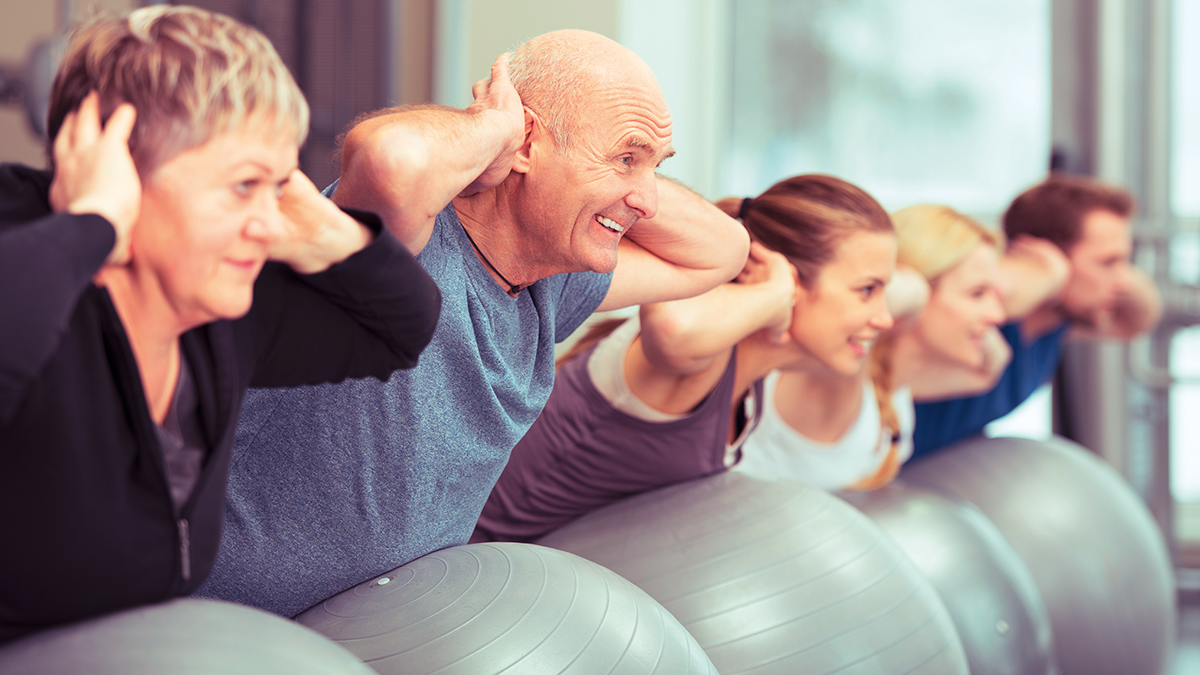Eine Gruppe aus 5 Personen unterschiedlichen Alters führt eine Fitnessübung jeweils auf einem Gymnastikball aus, im Vordergrund zwei Senioren