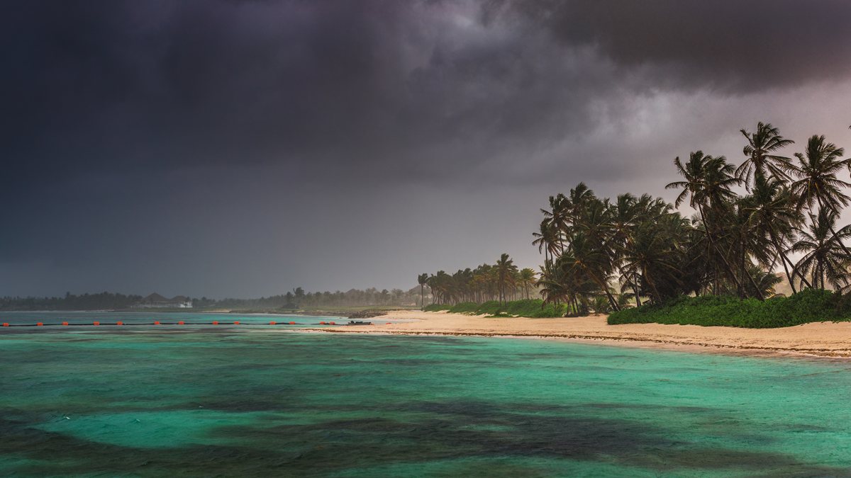 ESPRI, Projekte: Frühwarsystem Dominikanische Republik DOM Eine Insel mit Palmen und Strand, der Himmel ist mit dunklen Gewitterwolken verhangen.