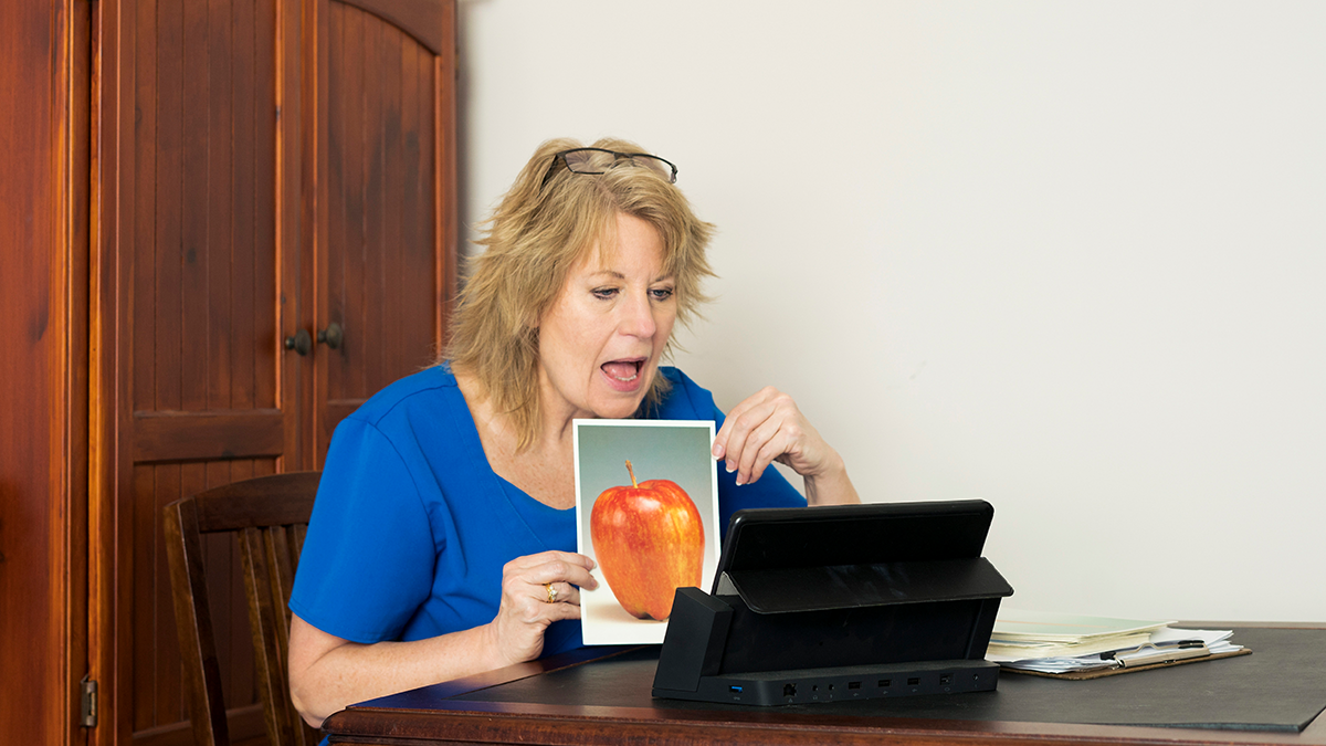 Blond female speech language pathologist, or speech therapist, wearing  royal blue scrubs provides teletherapy services by engaging with her patient online through her computer at home during the Covid-19 pandemic. Therapist is showing large single object pictures to elicit verbal responses that include naming for apraxia, word-finding for anomia, sentence descriptions for verbal expression.