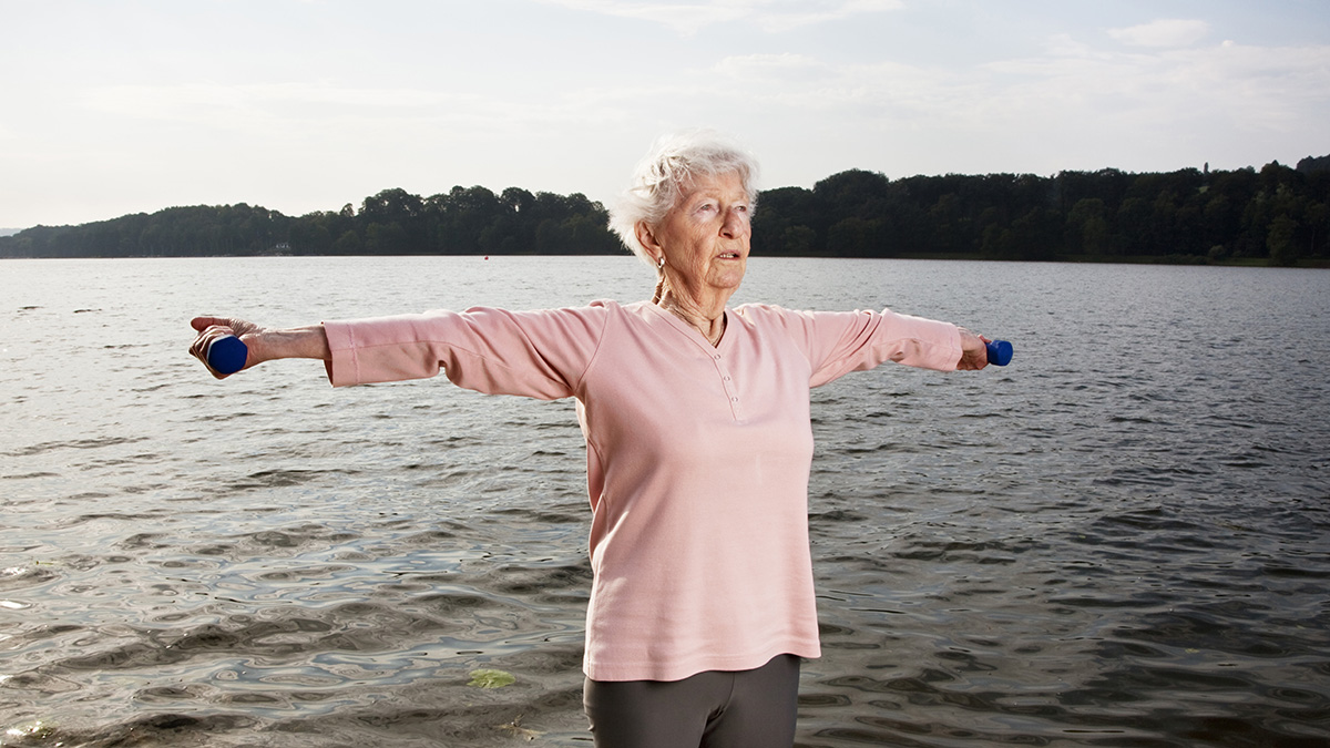 senior woman, 88 years old, very active and flexible, lifting dumbbells at a lake at a sunny summer morning