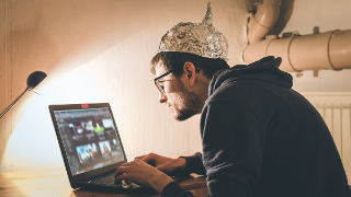 Mann mit Aluhut und Brille sitzt über einen Laptop gebeugt