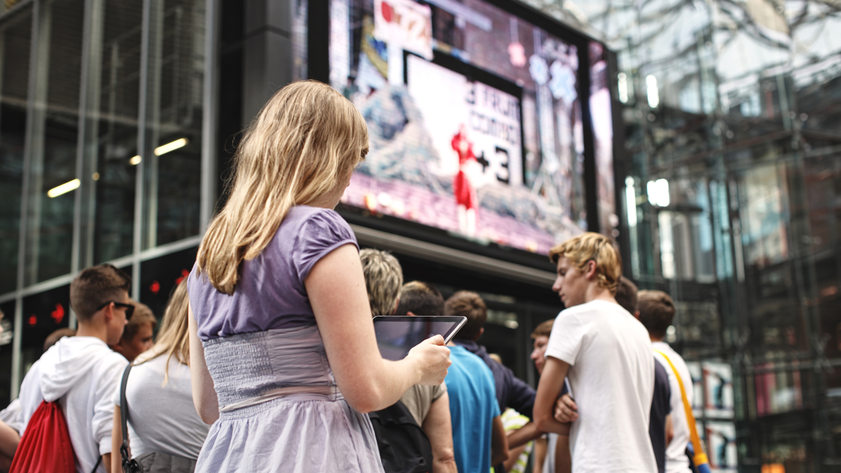 Junge Frau steht mit Tablet am Potsdamer Platz, im Hintergrund eine Schülergruppe und ein großer Werbescreen
