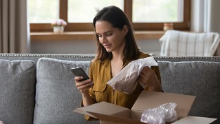 Eine Frau packt in ihrem Wohnzimmer einen Versandkarton aus und schaut dabei auf ihr Smartphone