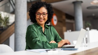 A professional woman wearing green works at a laptop in a stylish office environment. Bright and welcoming atmosphere emphasizes productivity and empowerment, with a focus on professional growth and positivity.