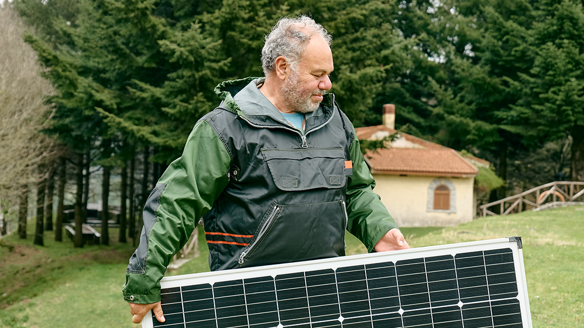 Middle aged bearded man carrying solar panel near house in rural zone. Alternative energy, renewable energy source and sustainable energy resources concept. Energy independence.