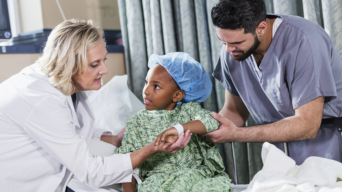 A 6 year old African-American girl sitting on a hospital gurney, with a doctor and nurse tending to her.  She is wearing a hospital gown and surgical cap. A nurse in scrubs, a young Middle Eastern man in his 20s, is standing on one side of her, and a female doctor, a mature woman in her 40s, is standing on the other side.