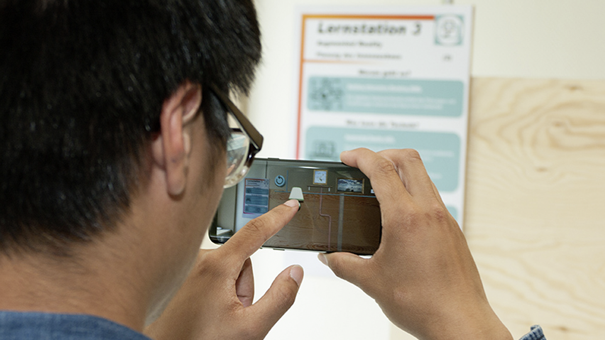 SQC, FAME, Projekte: DigiHand - Digitalisieung des Handwerks Ein Mann mit Brille hält ein Smartphone vor eine große Holzplatte. In der App auf dem Telefon bewegt er per AR sichtbare Objekte.