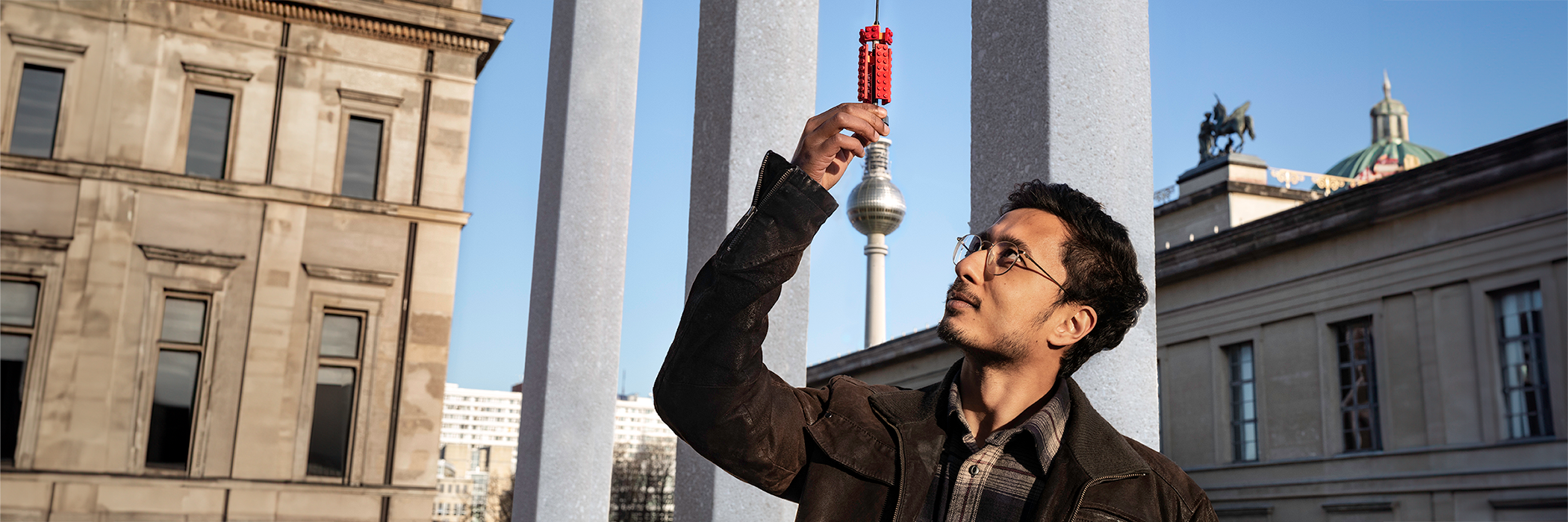 Young man with glasses holding up a lego antenna in front of the Berlin TV Tower