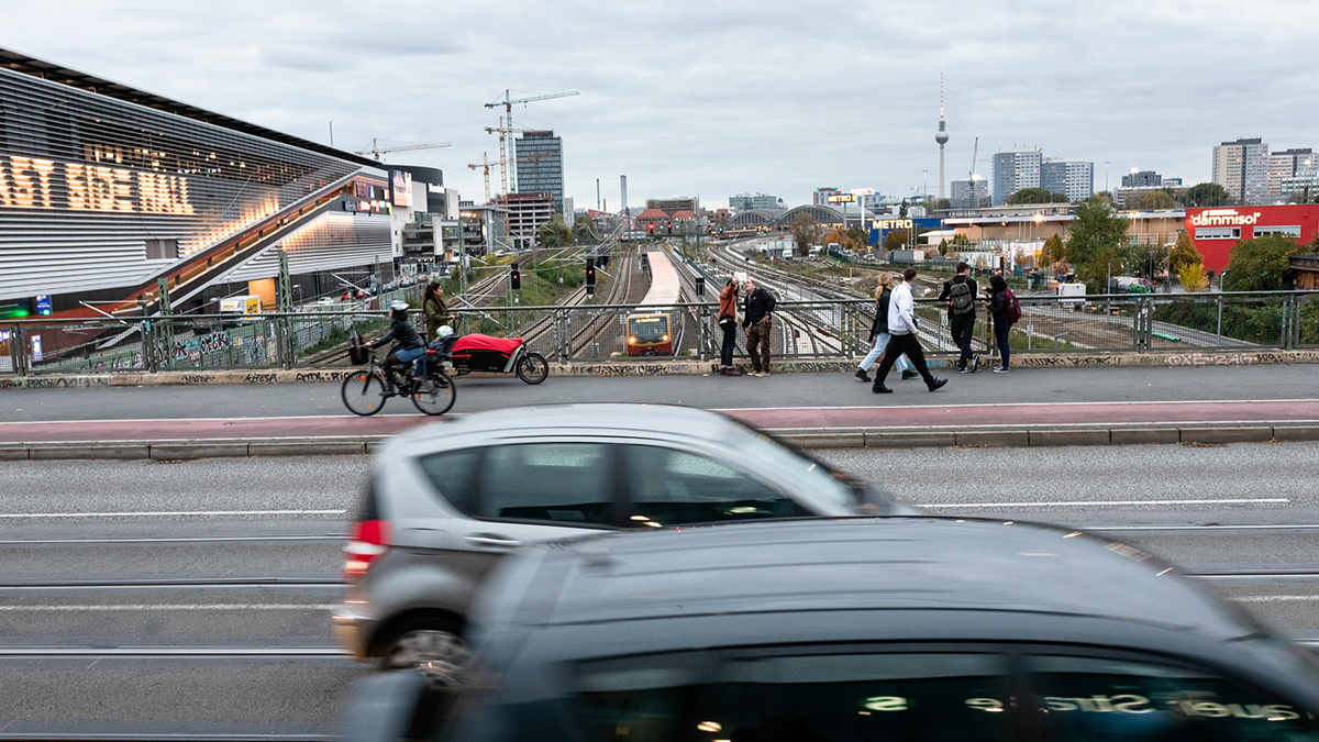 SQC, Themen: Urbane IKT, Smart Cities Brücke S-Bahnhof Warschauer-Straße, Berlin, mit Autos, Personen, im Hintergrund East Side Mall, eine S-Bahn auf den Gleisen und der Fernsehturm