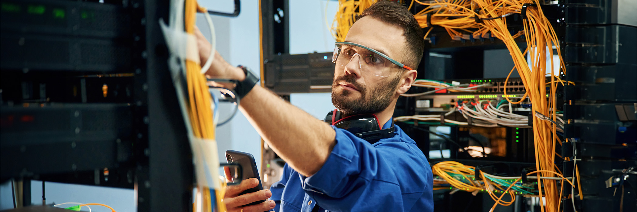 Holding smartphone. Young man is working with internet equipment and wires in server room.