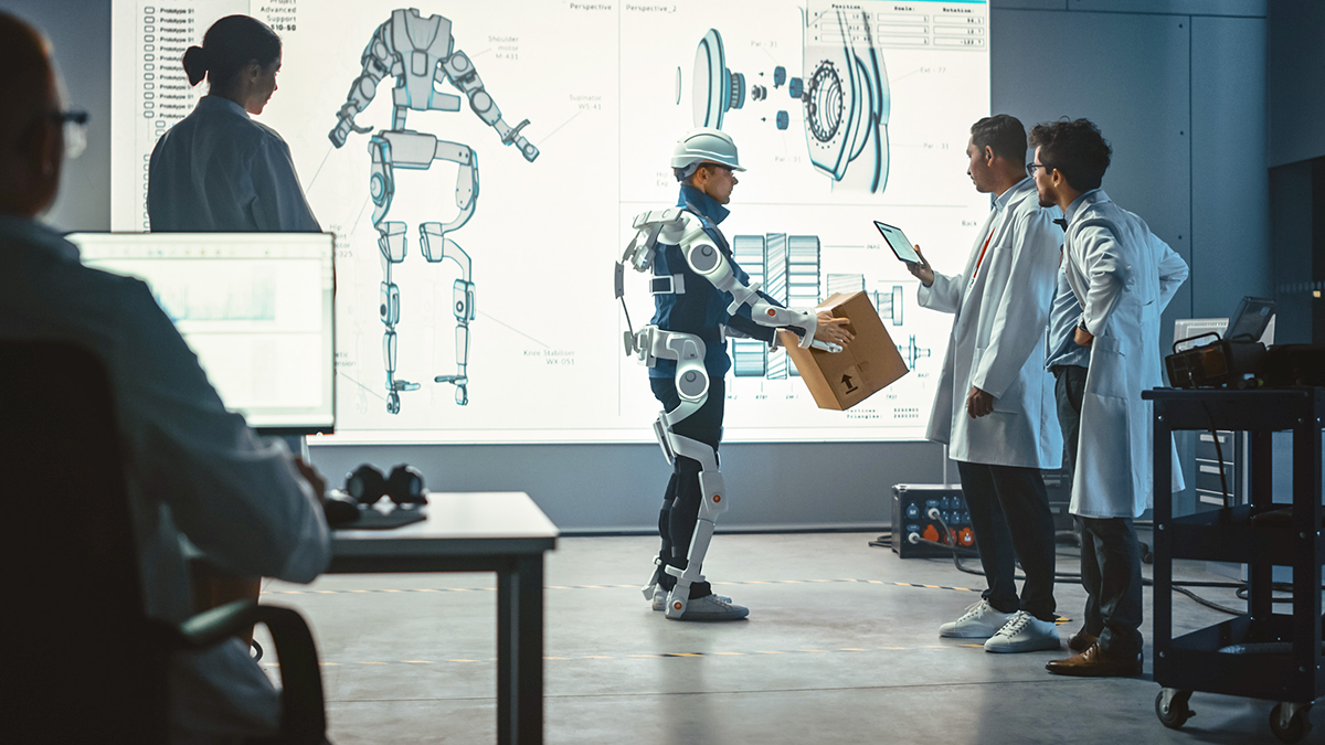 SQC, News: KI Standardisierung Im Entwicklungslabor: Ingenieure sehen sich den Prototyp eines robotischen Exoskeletts an - Präsentation mit einer Person, die es testet und eine schwere Pappschachtel hält. Entwurf eines tragbaren Exo-Anzugs zur Unterstützung von Lagerarbeitern