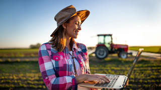 Female agronomist using laptop modern technology in agriculture. Young farmer standing in field in front of tractor machinery smiling.