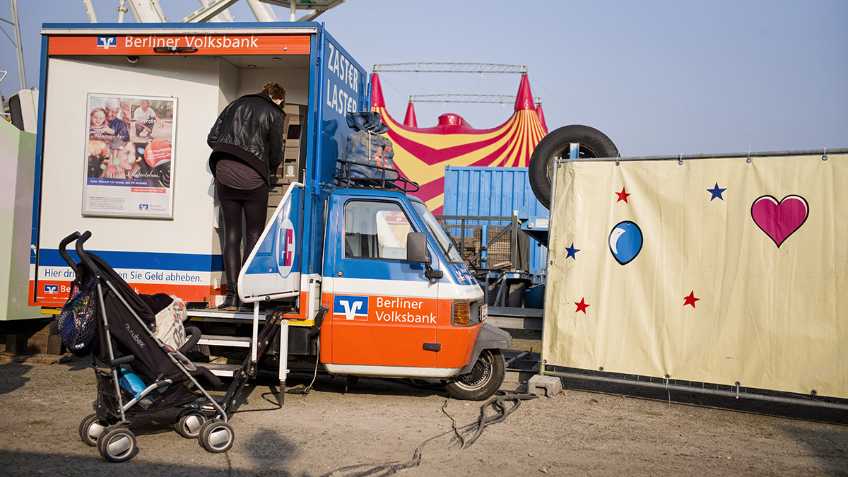 SQC, Projekte: DIAMONDS Mobiler Geldautomat auf einem Zirkusgelände, an dem eine Frau mit Kinderwagen Geld abhebt