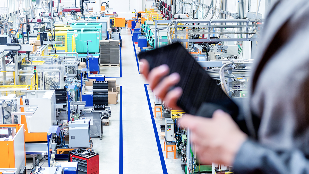 SQC, Projekte: IoTAC Horizontal color image of businesswoman - unrecognizable person - working with digital tablet in large futuristic factory. Woman standing on top of a balcony, holding touchpad and checking inventory of a manufacturing company on touchscreen tablet. Focus on futuristic machines in background, businesswoman's hands holding black tablet defocused.