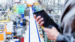 Horizontal color image of businesswoman - unrecognizable person - working with digital tablet in large futuristic factory. Woman standing on top of a balcony, holding touchpad and checking inventory of a manufacturing company on touchscreen tablet. Focus on futuristic machines in background, businesswoman's hands holding black tablet defocused.