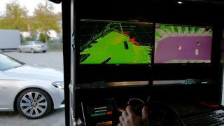 Aus einer mobilen Kontrollstation für Teleoperiertes Fahren mit 2 Bildschirmen und einem Lenkrad wird ein silbernes Fahrzeug gesteuert, dass im Hintergrund zu sehne ist.