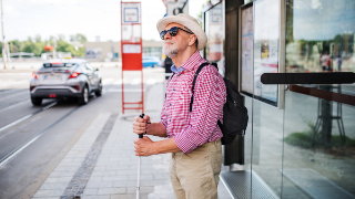 Ein blinder Mann mit Hut, Sonnenbrille und Langstock steht an einer Bushaltestelle.