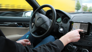 Person sitzt in einem stehenden Auto hinter dem Lankrad und bedient mit der rechten Hand ein Navigationspad auf dem die TEAM App läuft.