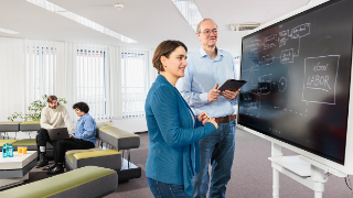 Eine Wissenschaftlerin und ein Wissenschaftler stehen an einem Whiteboard auf dem ein Verlaufsdiagramm für das eGovernment Labor abgebildet ist. Im Hintergrund sitzen zwei weitere Wissenschaftliche Mitarbeitende auf einem grünen Sofa, die gemeinsam an einem Laptop arbeiten.