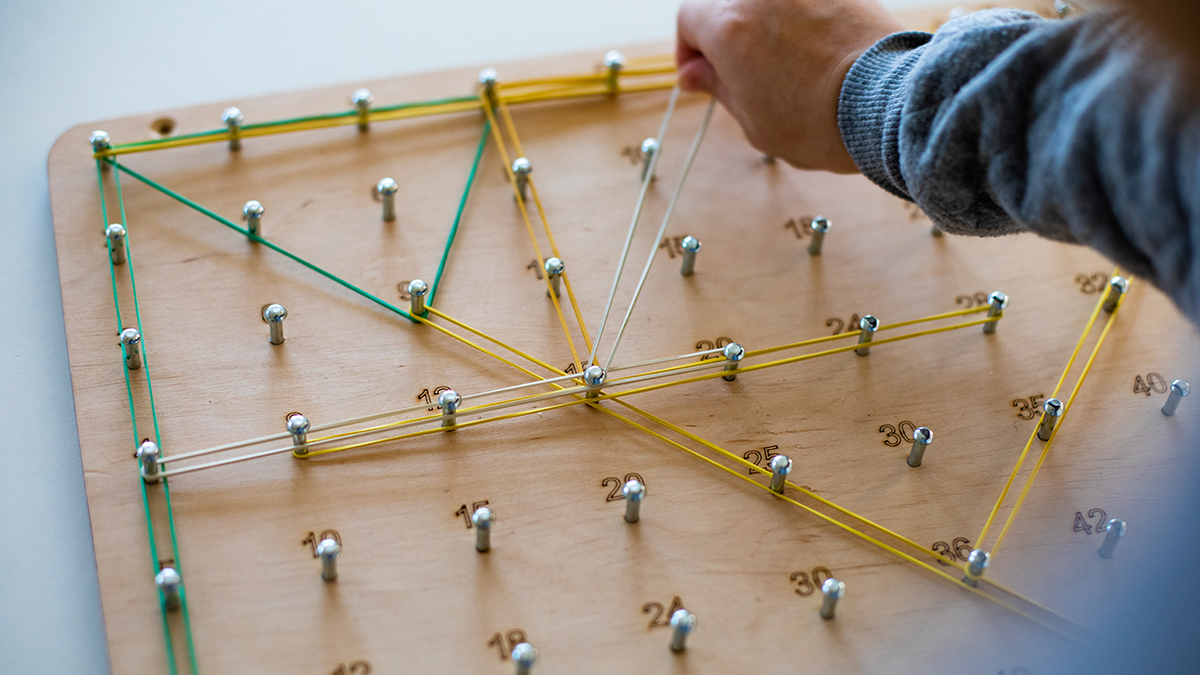 Geoboard for developing mathematical thinking and problem solving skills: Closeup cropped child learning to use geoboard. Wooden board with rows of half driven nails with numbers and rubber bands. Top view, selective focus.