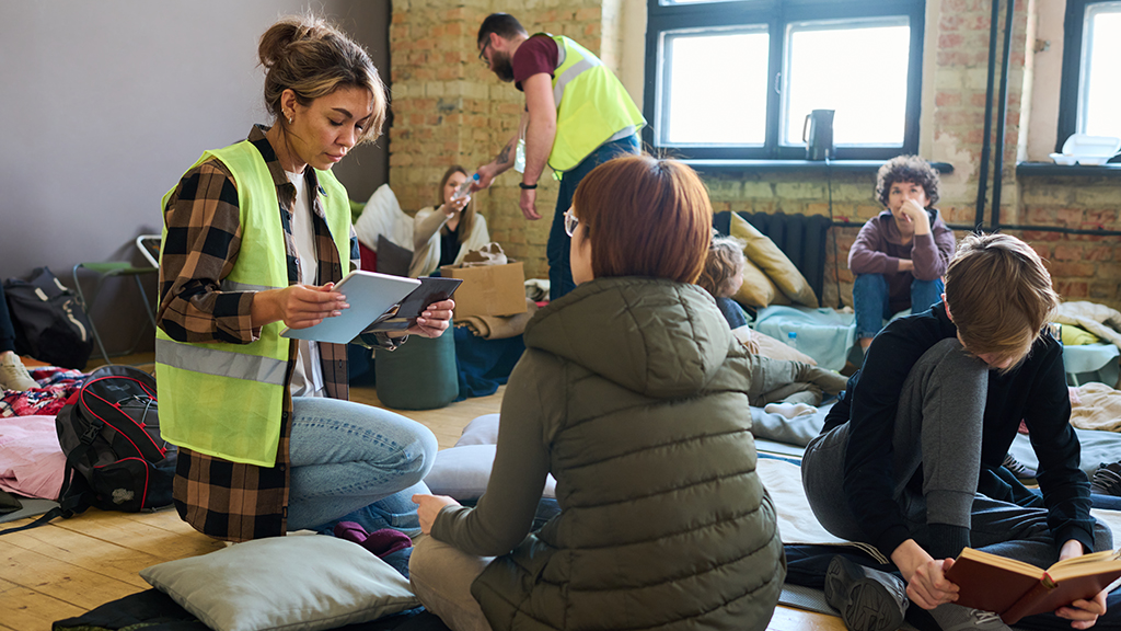 Female volunteer with mobile gadgets sitting on squats in front of refugee sitting on her sleeping place in temporary shelter