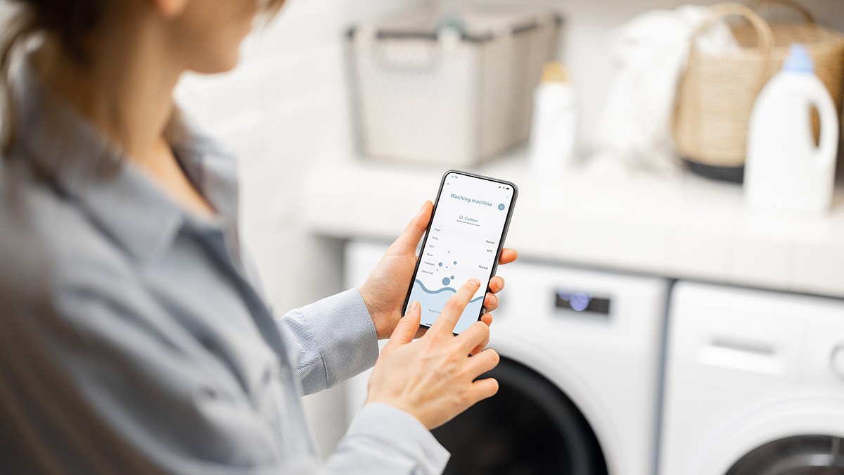 Woman controls washing machine with a smartphone, holding phone with a running program for smart home appliances