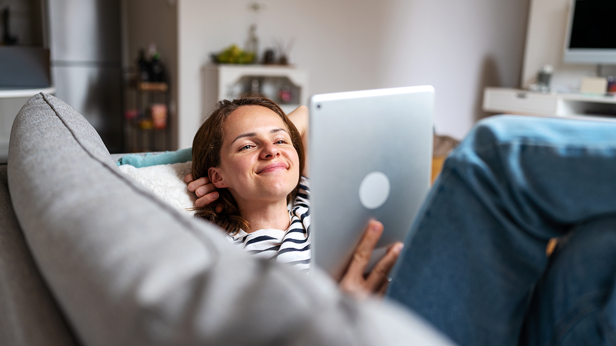 Woman watching vlog on internet and smiling while lying on coach at home